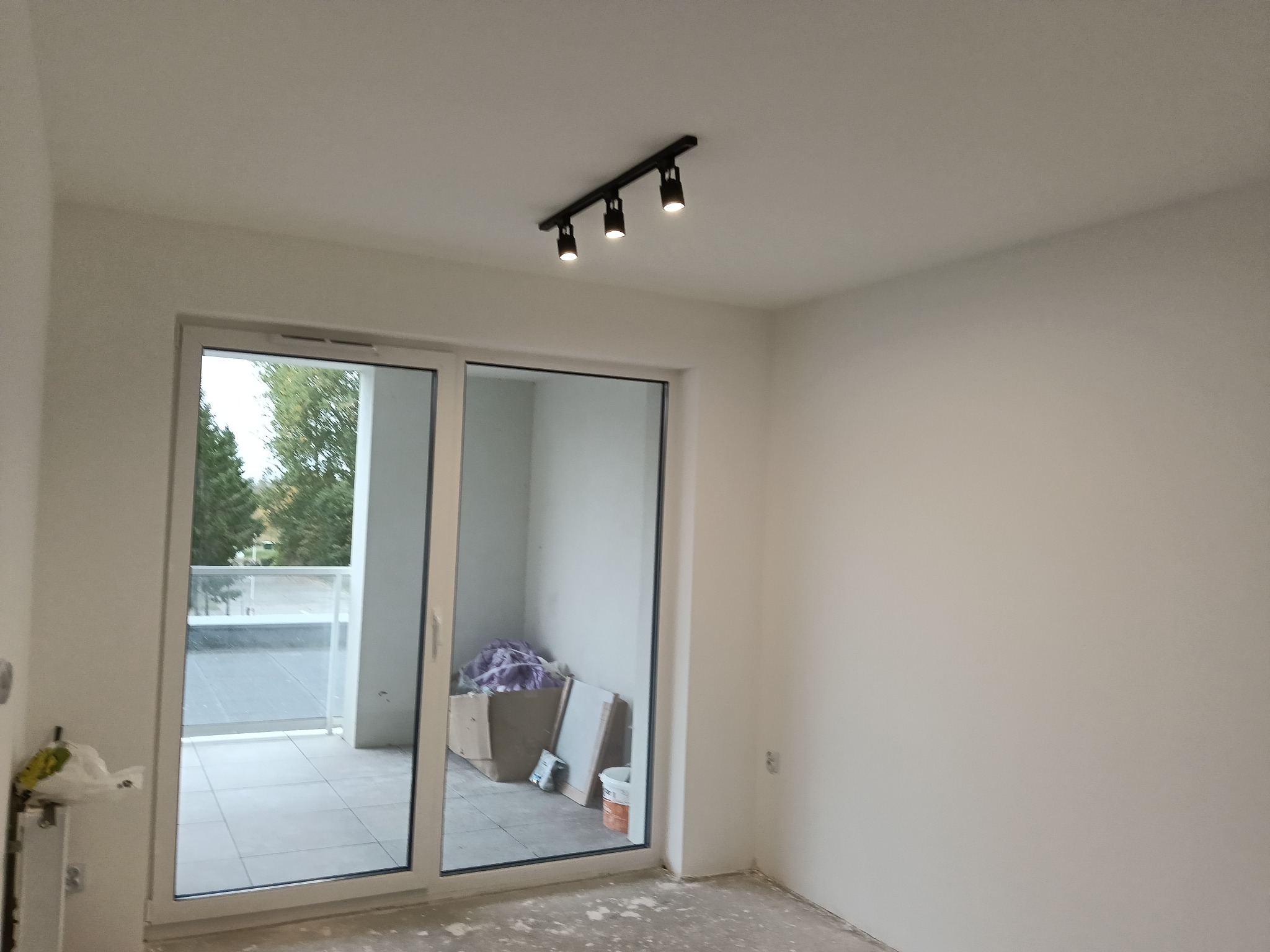 Jasne wnętrze z dużym oknem balkonowym i nowoczesnym oświetleniem sufitowym. Ściany świeżo pomalowane na biało, podłoga przygotowana do dalszych prac wykończeniowych.