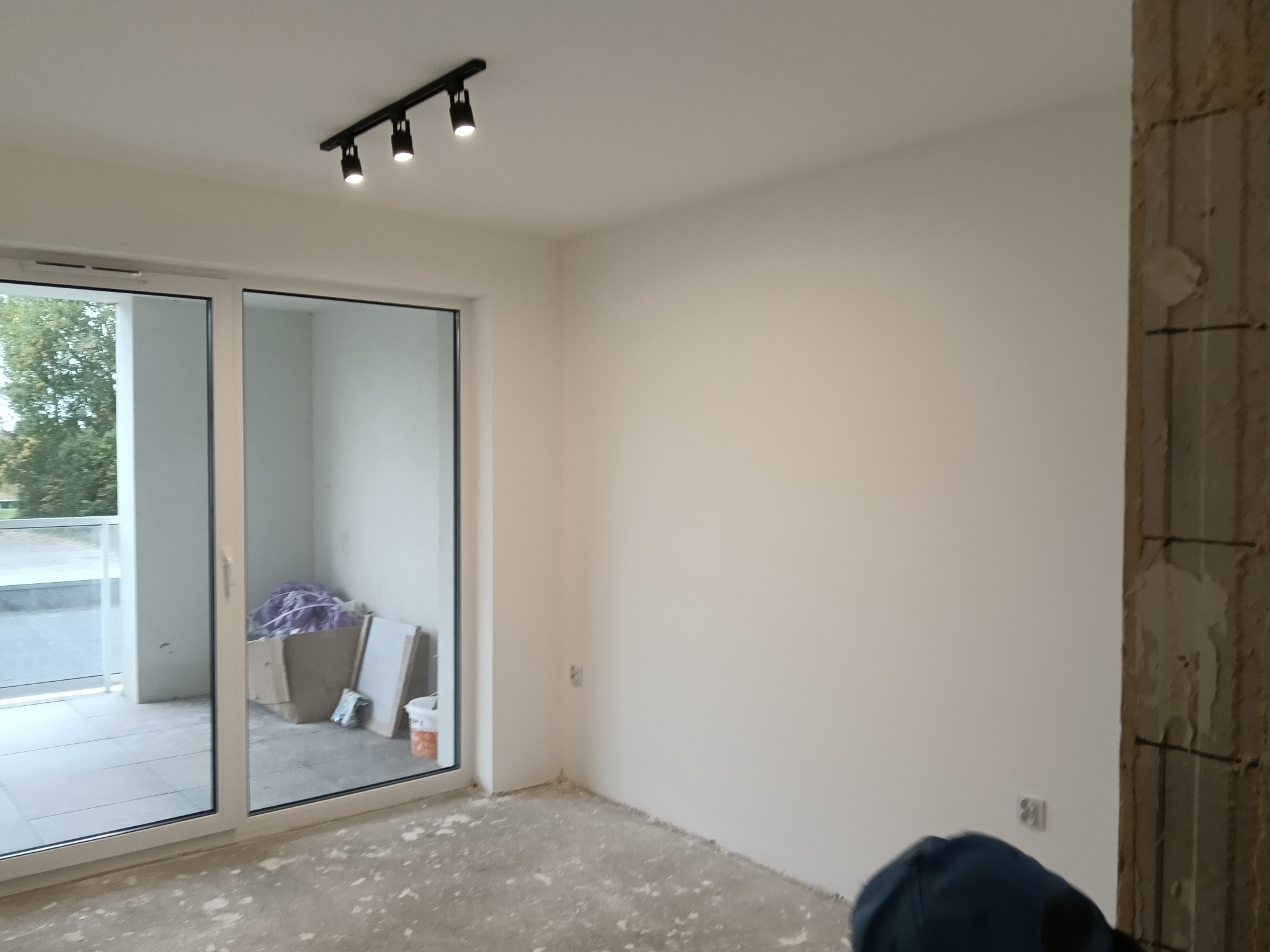 Jasne wnętrze po remoncie: gładkie, białe ściany, surowe wykończenie podłogi, przesuwne drzwi balkonowe i nowoczesne oświetlenie punktowe na suficie. Widoczny fragment ściany w stanie surowym.