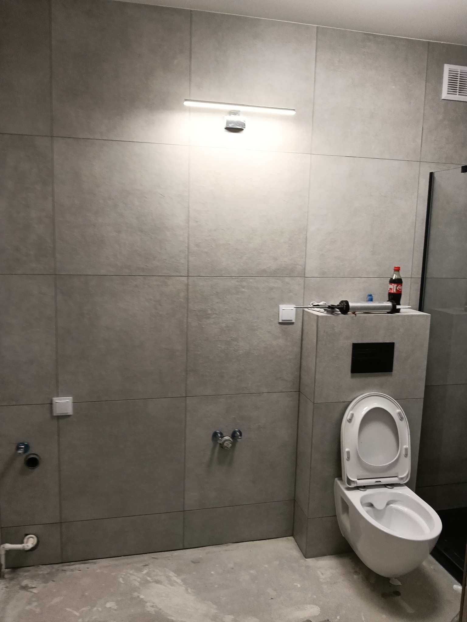 Szara łazienka w trakcie wykańczania: zamontowana toaleta wisząca, płytki ścienne, narzędzia na obudowie stelaża, przygotowane przyłącza wodne i kanalizacyjne.