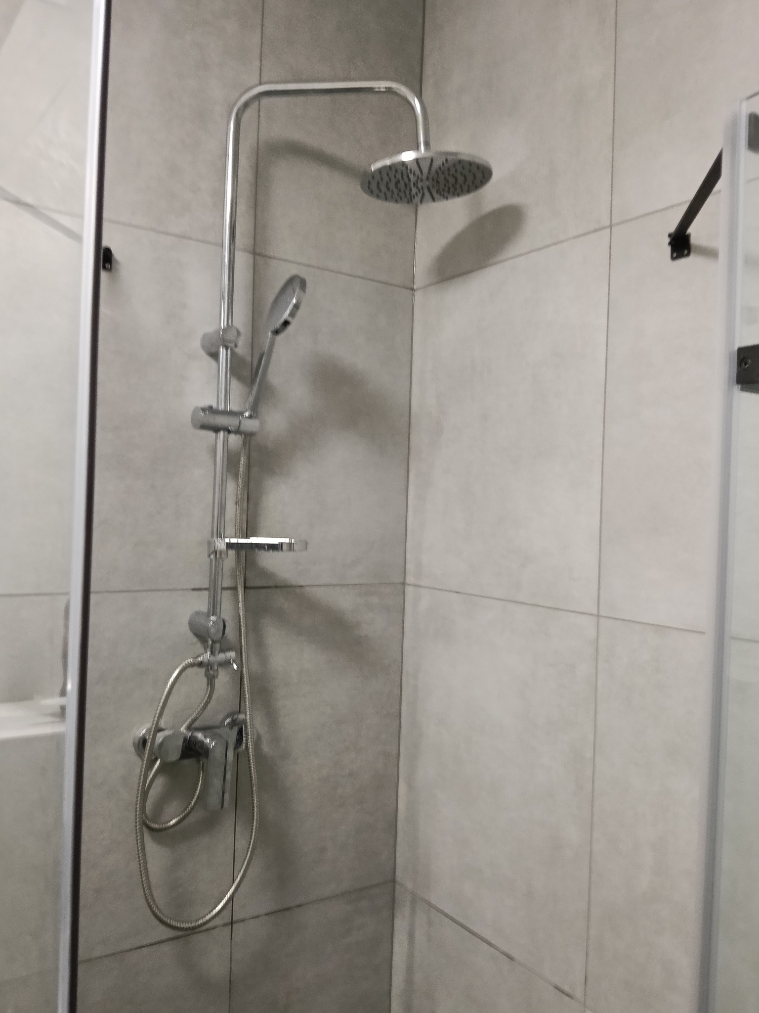 Nowoczesny zestaw prysznicowy z deszczownicą i słuchawką, zamontowany na jasnoszarych płytkach w kabinie. Minimalistyczny design, widoczne elementy mocujące.