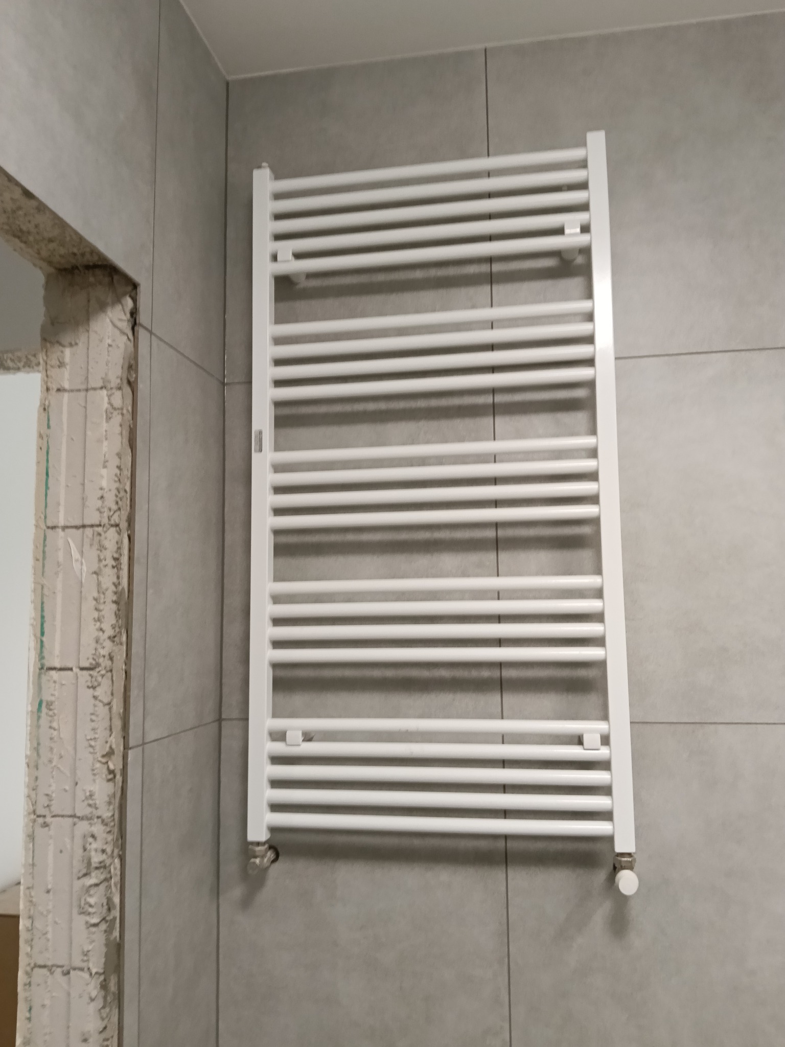 Biały grzejnik łazienkowy na szarej ścianie z płytek, widoczny fragment surowej ściany po lewej stronie, sugerujący remont łazienki.