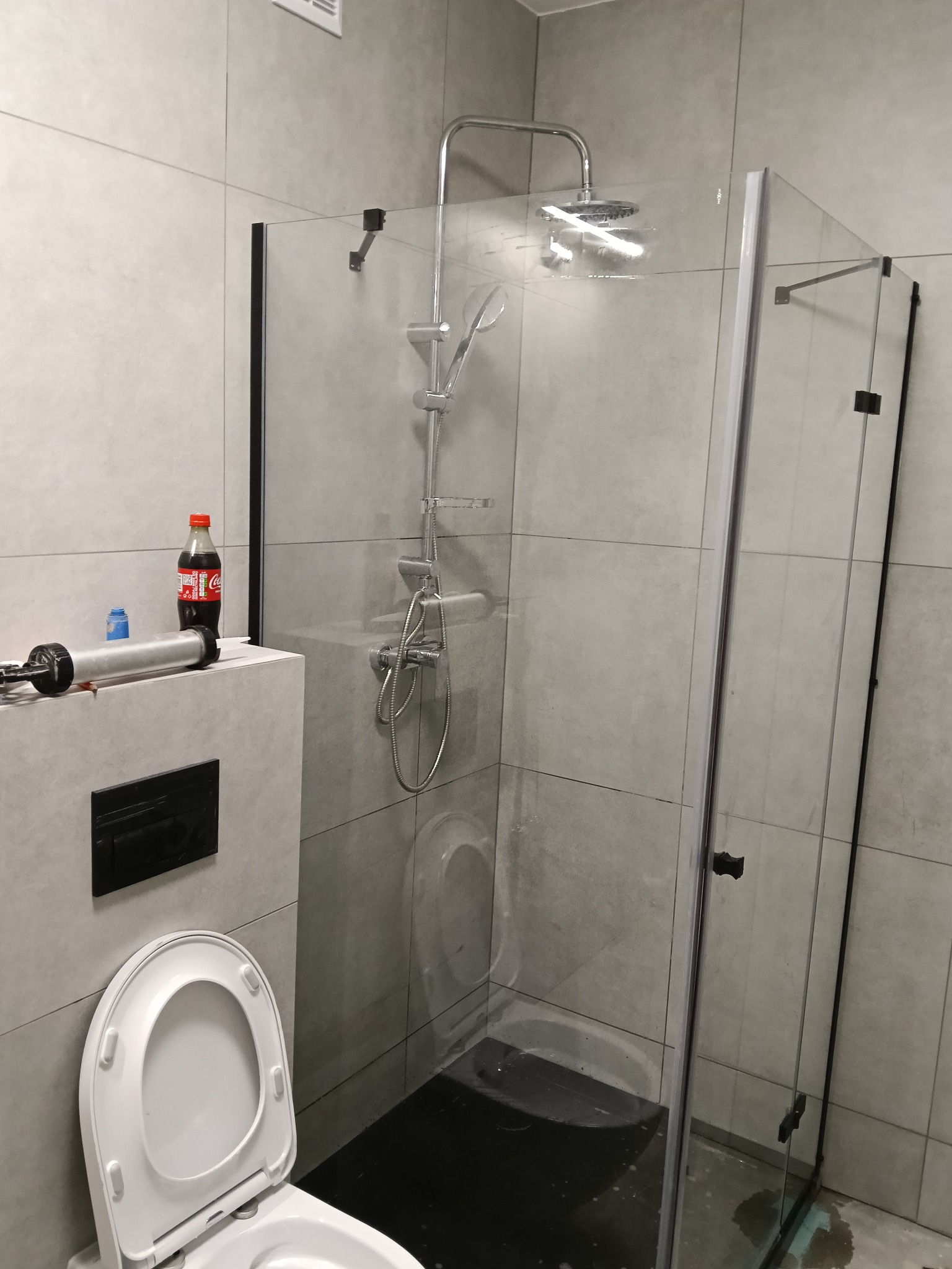 Nowoczesna kabina prysznicowa z czarnymi profilami w łazience wykończonej szarymi płytkami. Widoczny silikon i narzędzia, sugerujące montaż lub uszczelnianie.