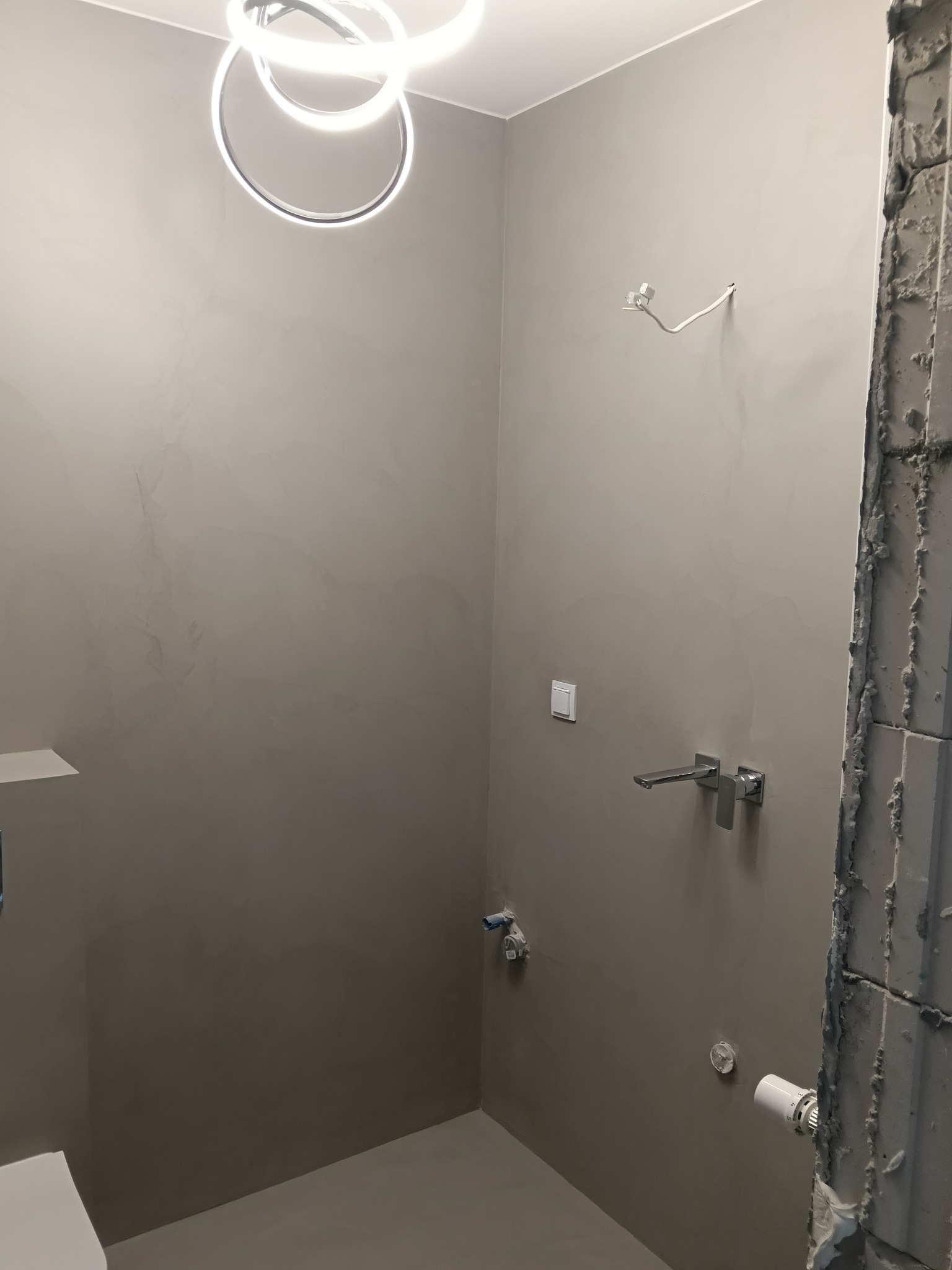 Minimalistyczna łazienka w szarościach z nowoczesnym oświetleniem sufitowym. Widoczne elementy instalacji wodnej i fragment ściany w trakcie prac wykończeniowych.