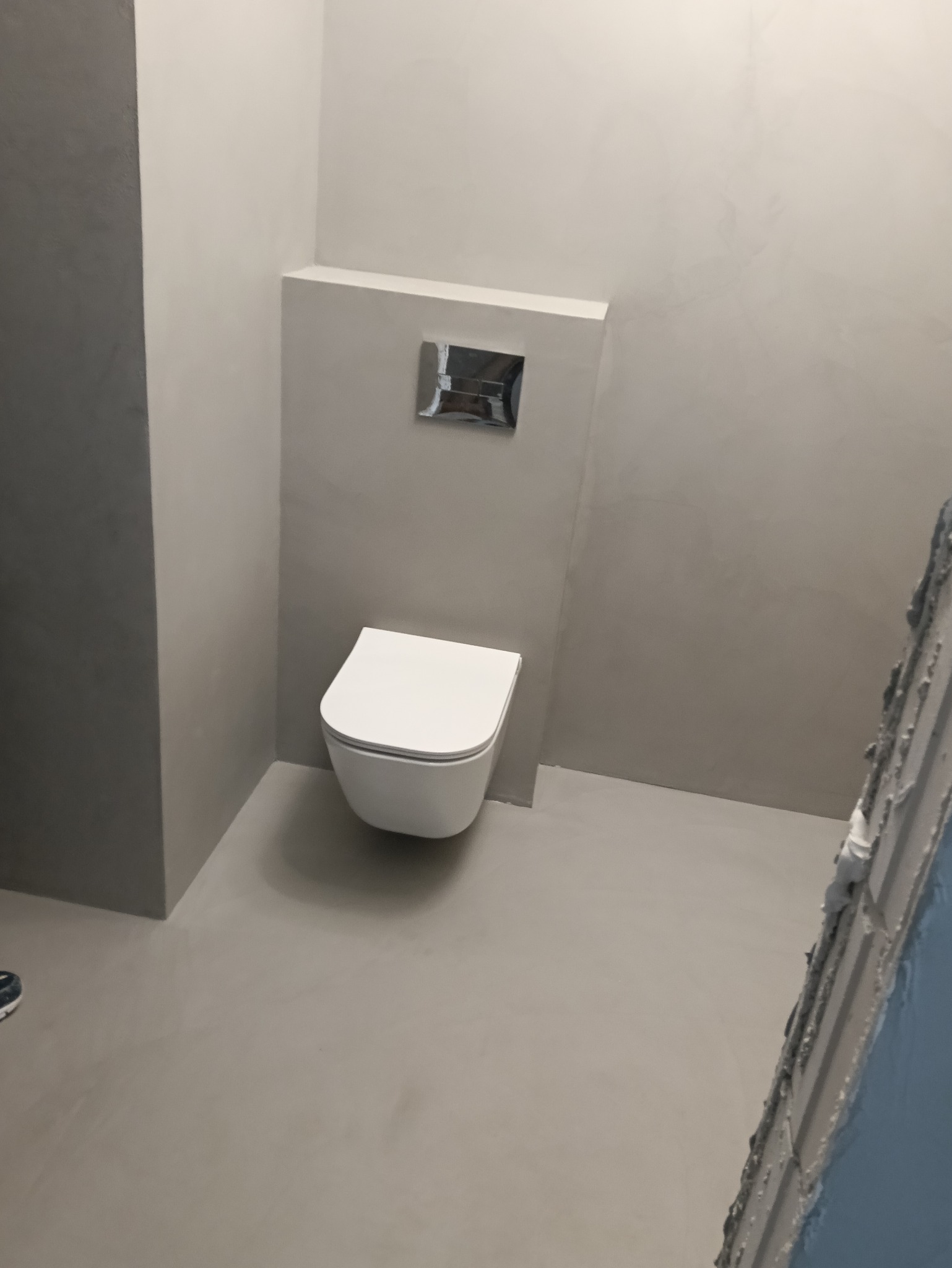 Minimalistyczna toaleta podwieszana w wykończonej na szaro łazience. Widoczne gładkie ściany i podłoga w jednolitej tonacji, chromowany przycisk spłukiwania.