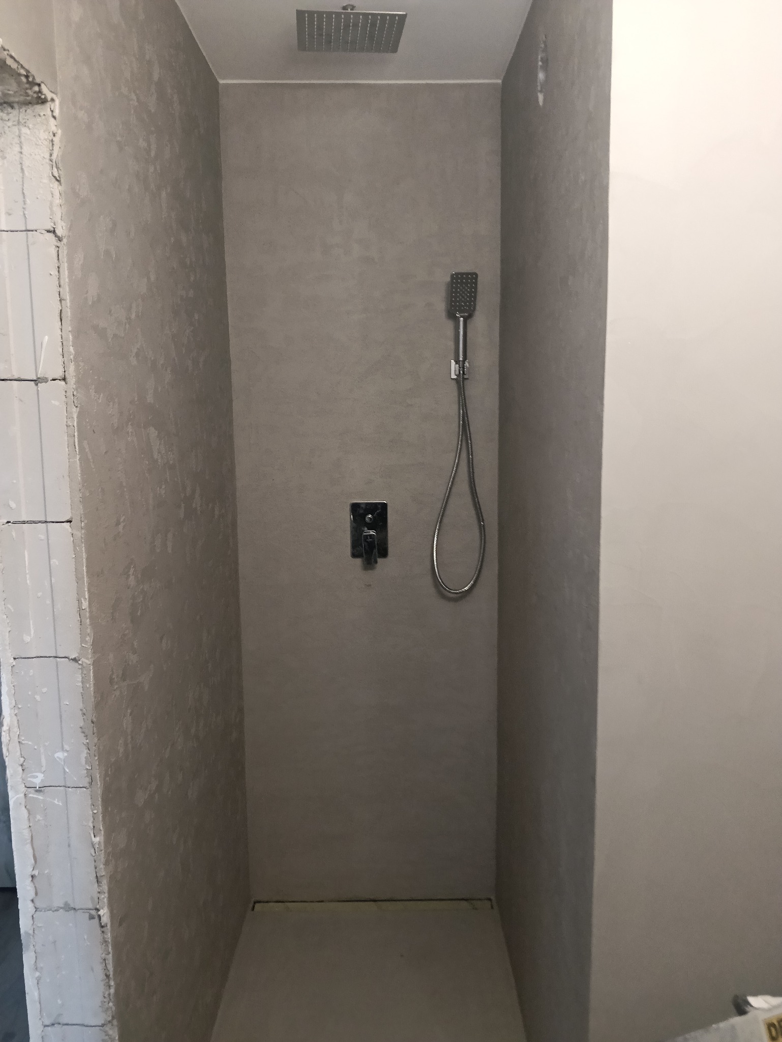 Wykończona kabina prysznicowa z szarą, teksturowaną ścianą, deszczownicą i baterią. Fragment ściany z surowego muru po lewej stronie. Minimalistyczny design.