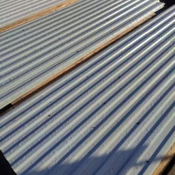 Stos blachy trapezowej w kolorze szarym z widocznymi kroplami wody, ułożony na drewnianych legarach pod lekkim kątem.