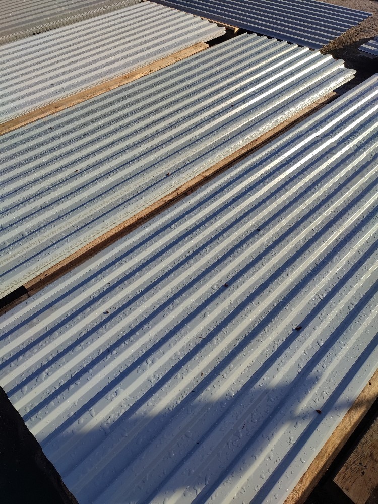 Stos blachy trapezowej w kolorze szarym z widocznymi kroplami wody, ułożony na drewnianych legarach pod lekkim kątem.