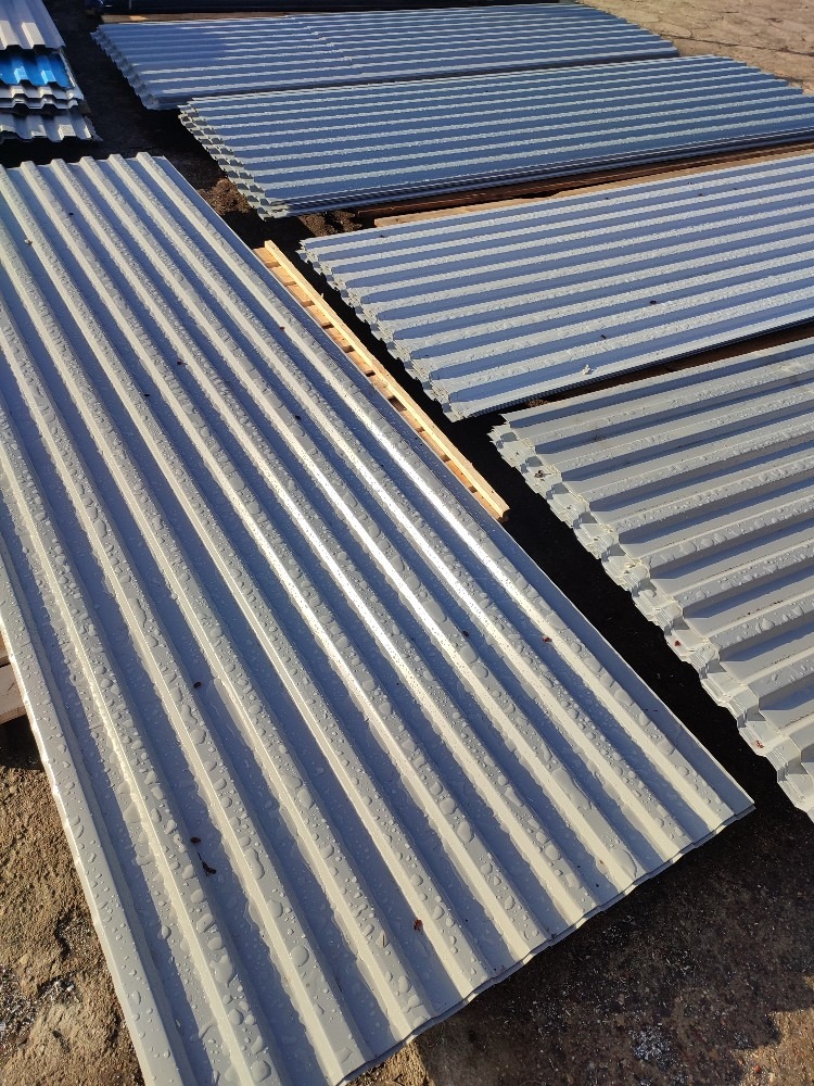 Stos arkuszy blachy falistej w odcieniach szarości, ułożonych na drewnianych legarach, z widocznymi kroplami wody na powierzchni i ziemistym tłem.