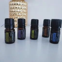 Rząd pięciu buteleczek olejków eterycznych marki dōTERRA: Oregano, Lavender, Lemon, Frankincense i Deep Blue, ustawionych na białym blacie, z dekoracyjną, złotą lampą w tle.