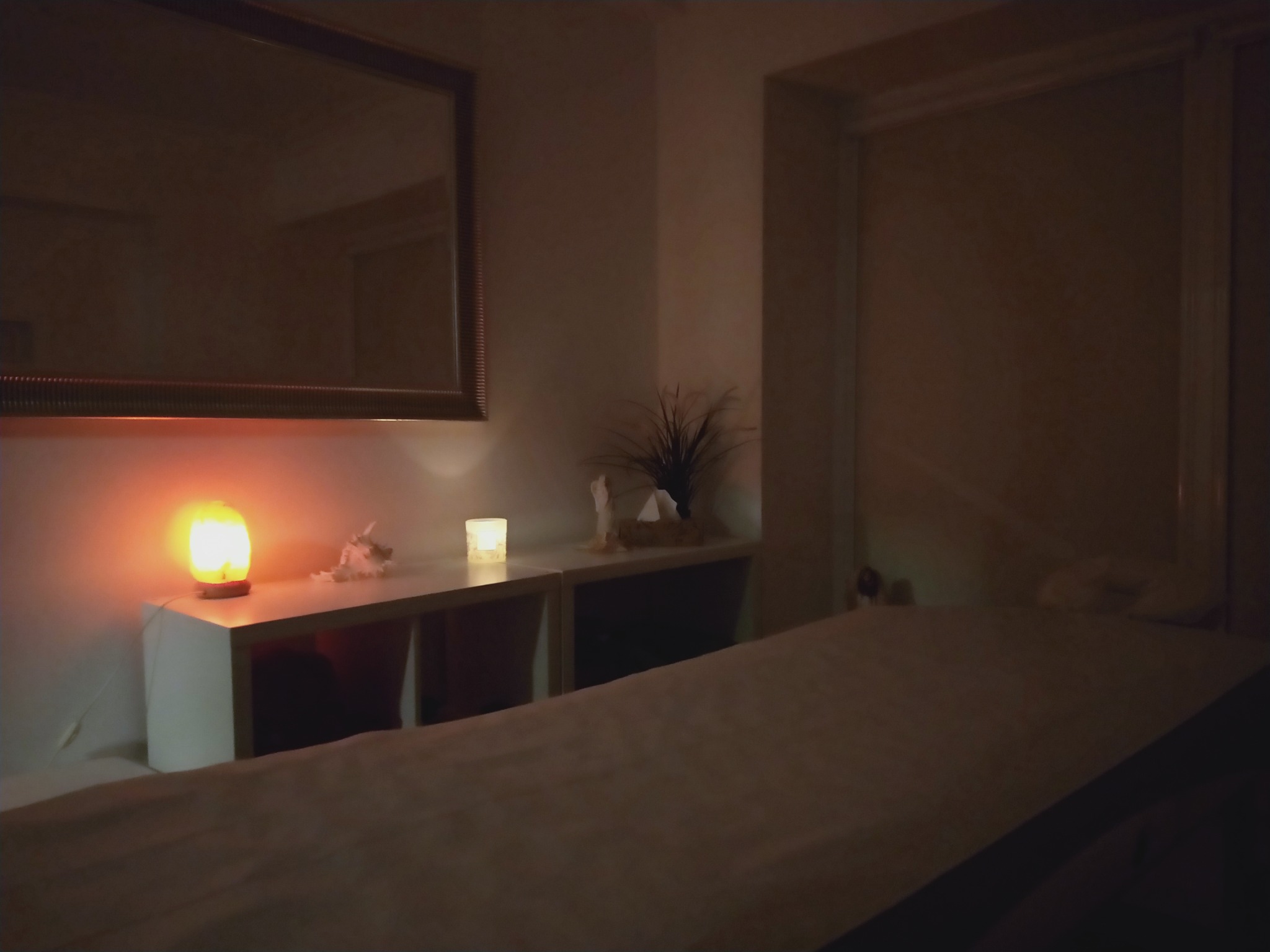 Przyciemnione wnętrze gabinetu z łóżkiem do masażu, lampką solną na szafce nocnej i lustrem na ścianie.