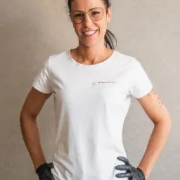 Uśmiechnięta kobieta w okularach i czarnych rękawicach ochronnych, ubrana w białą koszulkę z logo 'Czysta Sprawa', pozuje na tle szarej ściany.