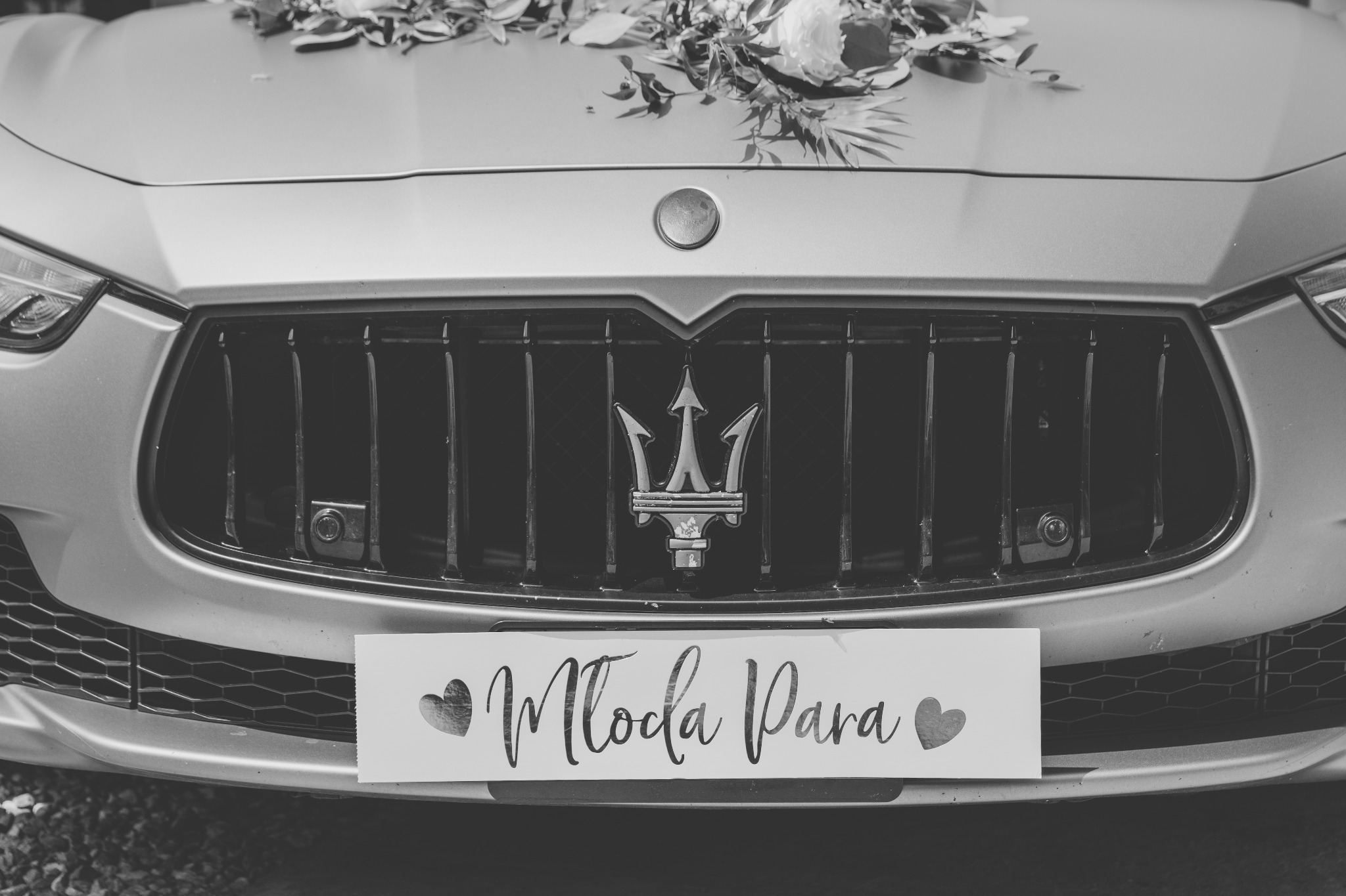 Czarno-białe zdjęcie przodu Maserati z dekoracją kwiatową na masce i tabliczką 'Młoda Para' na grillu.