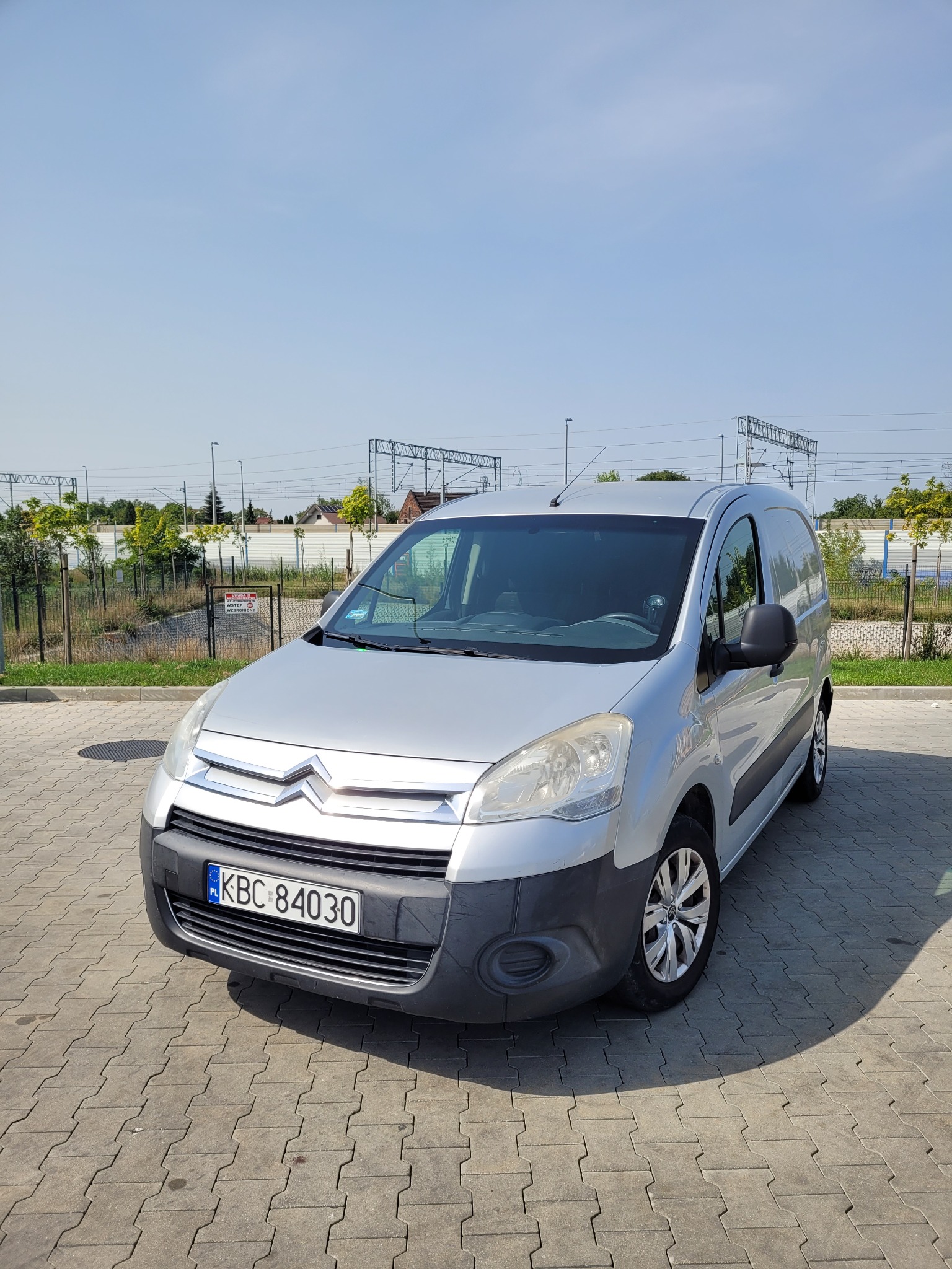 Srebrny samochód dostawczy Citroen Berlingo zaparkowany na kostce brukowej, z widoczną tablicą rejestracyjną KBC 84030, tle słupów trakcji kolejowej i zieleni.