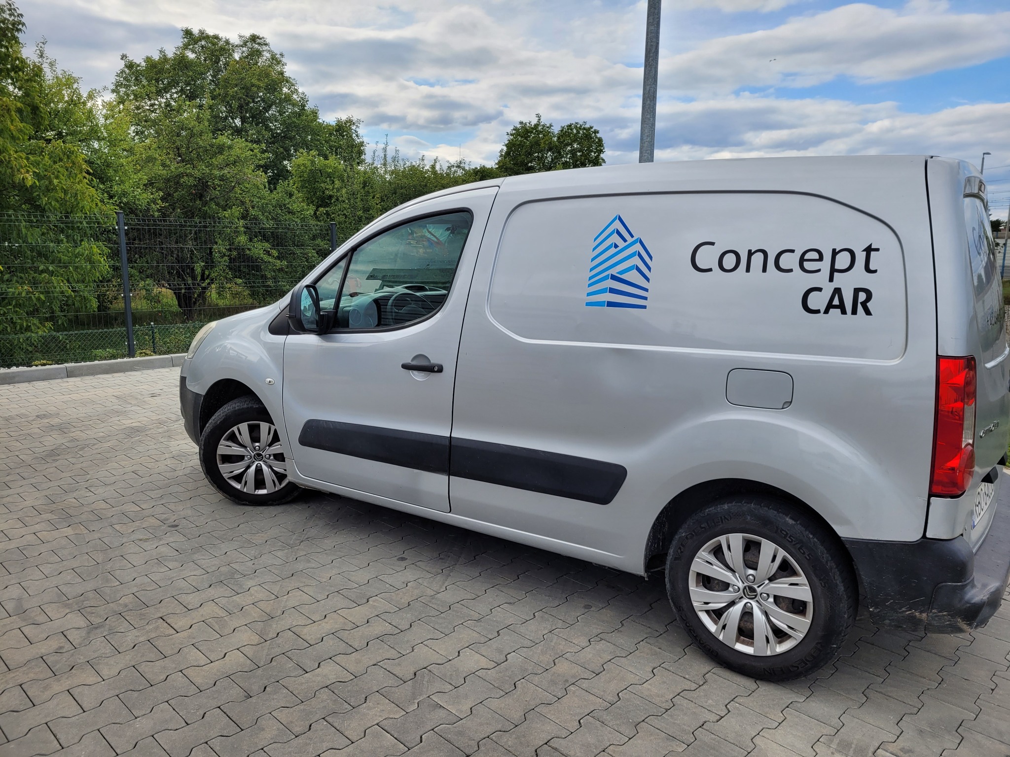 Srebrny samochód dostawczy z niebieskim logo i napisem 'Concept CAR' na tle kostki brukowej i zieleni.