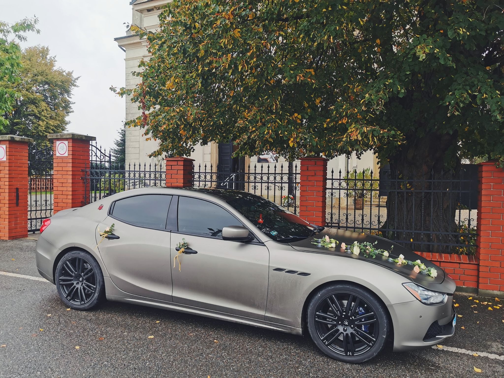 Elegancki, matowy, szary samochód Maserati udekorowany kwiatami na masce i klamkach, zaparkowany przed budynkiem z cegły i kutym ogrodzeniem.