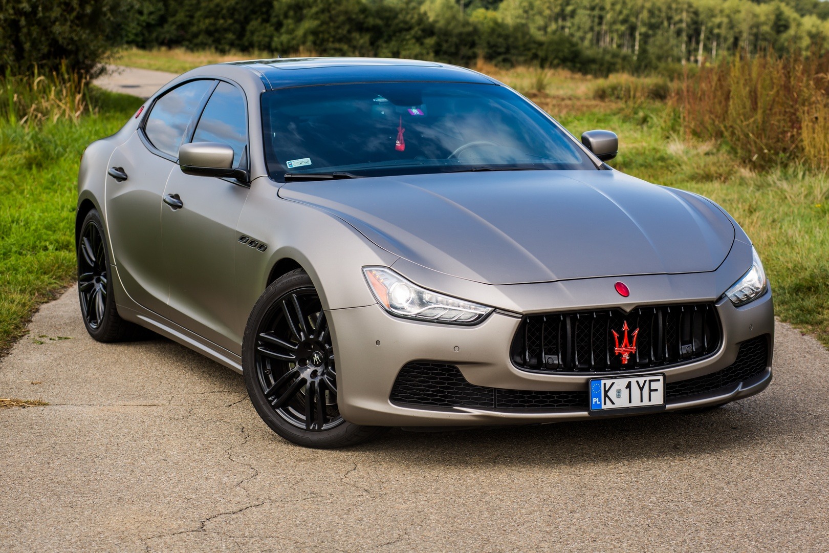 Matowy, szary Maserati Ghibli z czarnymi felgami, zaparkowany na asfalcie z widocznym krajobrazem polnym w tle.