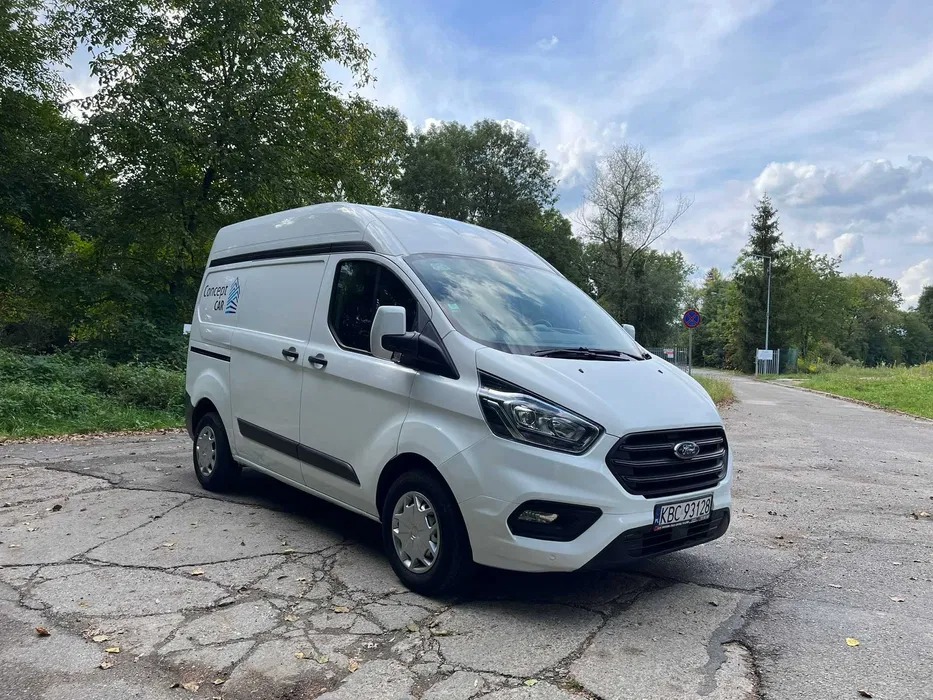 Biały furgon Ford Transit z wysokim dachem i logo firmy Concept CAR na boku, zaparkowany na popękanej betonowej nawierzchni z widoczną tablicą rejestracyjną KBC 93128, w tle zieleń drzew i fragment...