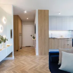 Nowoczesne wnętrze mieszkania z drewnianą podłogą w jodełkę, białymi szafkami z okrągłymi lampami na ścianie, drewnianymi szafami w korytarzu i kuchnią z drewnianymi szafkami oraz niebieską kanapą...