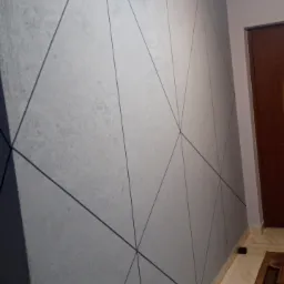 Szara ściana wewnętrzna z geometrycznym wzorem czarnych linii tworzących trójkąty, widoczna podłoga z płytek i fragment drzwi.