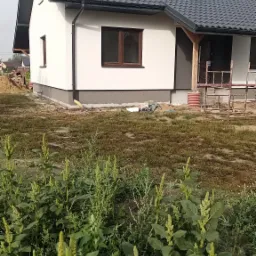 Ocieplony dom jednorodzinny z szarą dachówką, brązowymi oknami i fragmentem rusztowania w tle, otoczony wysokimi chwastami.