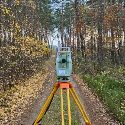 Zielono-biały teodolit na żółtym statywie stoi na polnej drodze w lesie jesienią, widok z przodu.