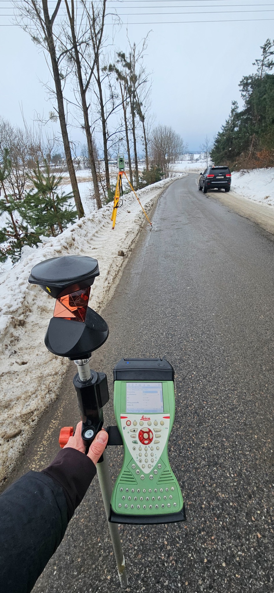 Pomiar geodezyjny w terenie zimowym: ręka trzyma tyczkę z pryzmatem i kontrolerem Leica, w tle droga, samochód i sprzęt na statywie.