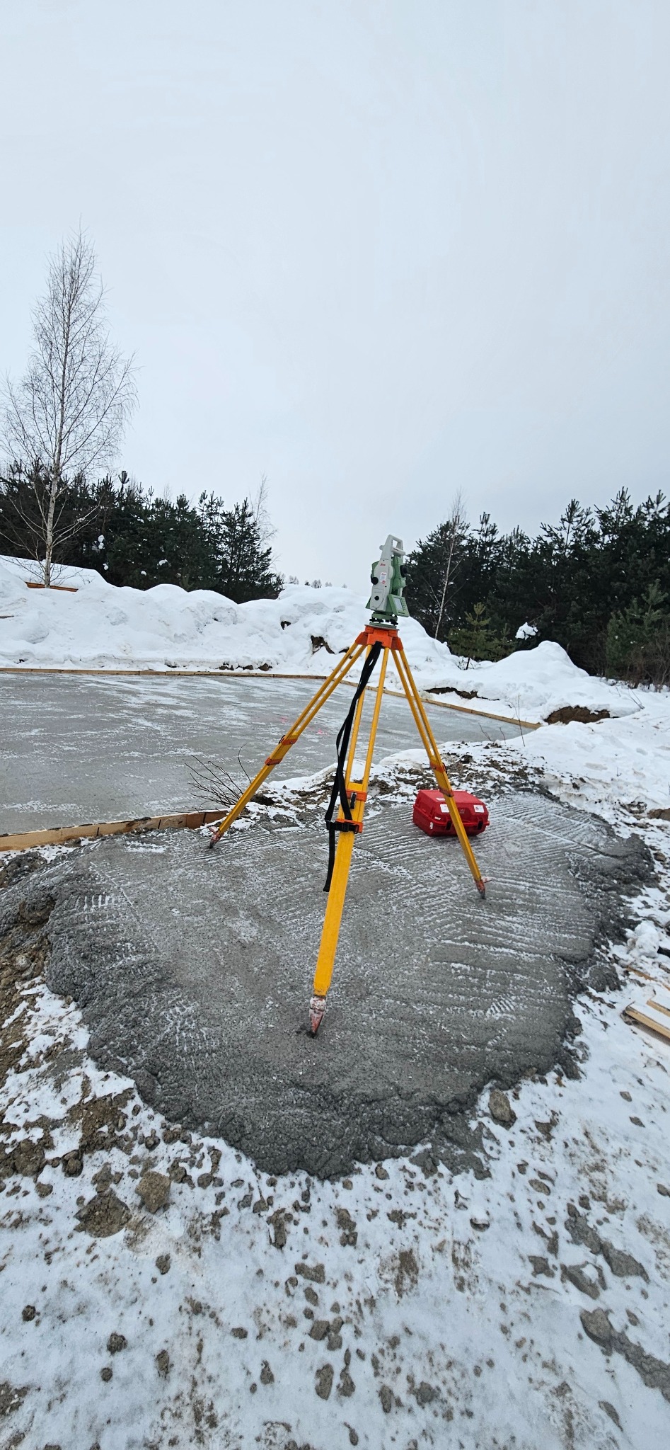 Żółty niwelator na trójnogu ustawiony na świeżym fundamencie w zimowym krajobrazie. Widoczny czerwony futerał i zasypany śniegiem teren.