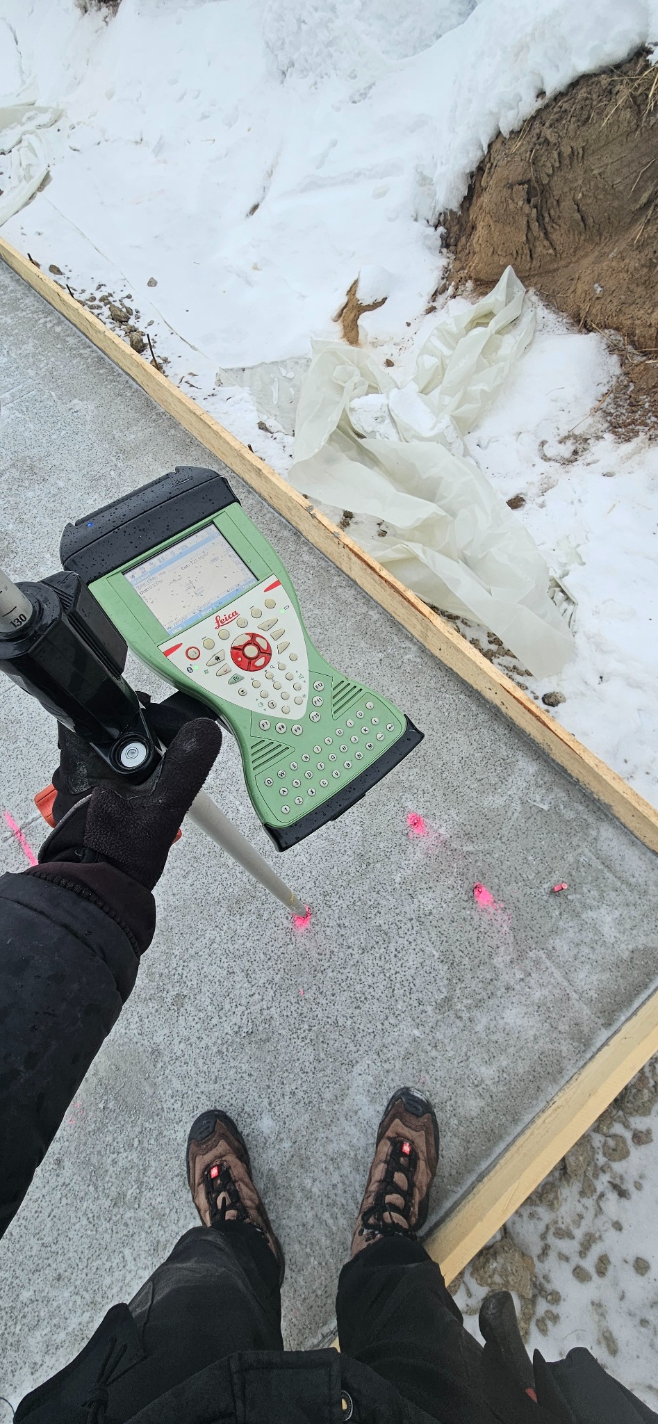 Pomiar geodezyjny Leica na betonowej płycie fundamentowej z drewnianym szalunkiem, widoczne punkty laserowe i śnieg w tle. Praca w toku.
