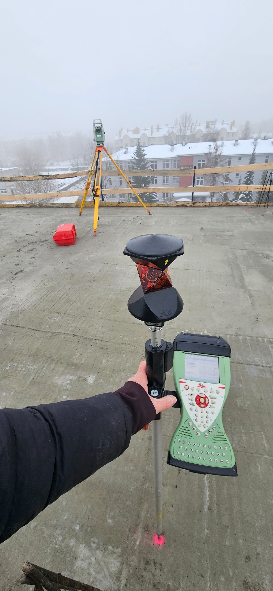 Praca z tachimetrem Leica na budowie w zimowy dzień. W tle budynki w lekkiej mgle. Czerwony punkt lasera na szarej powierzchni.