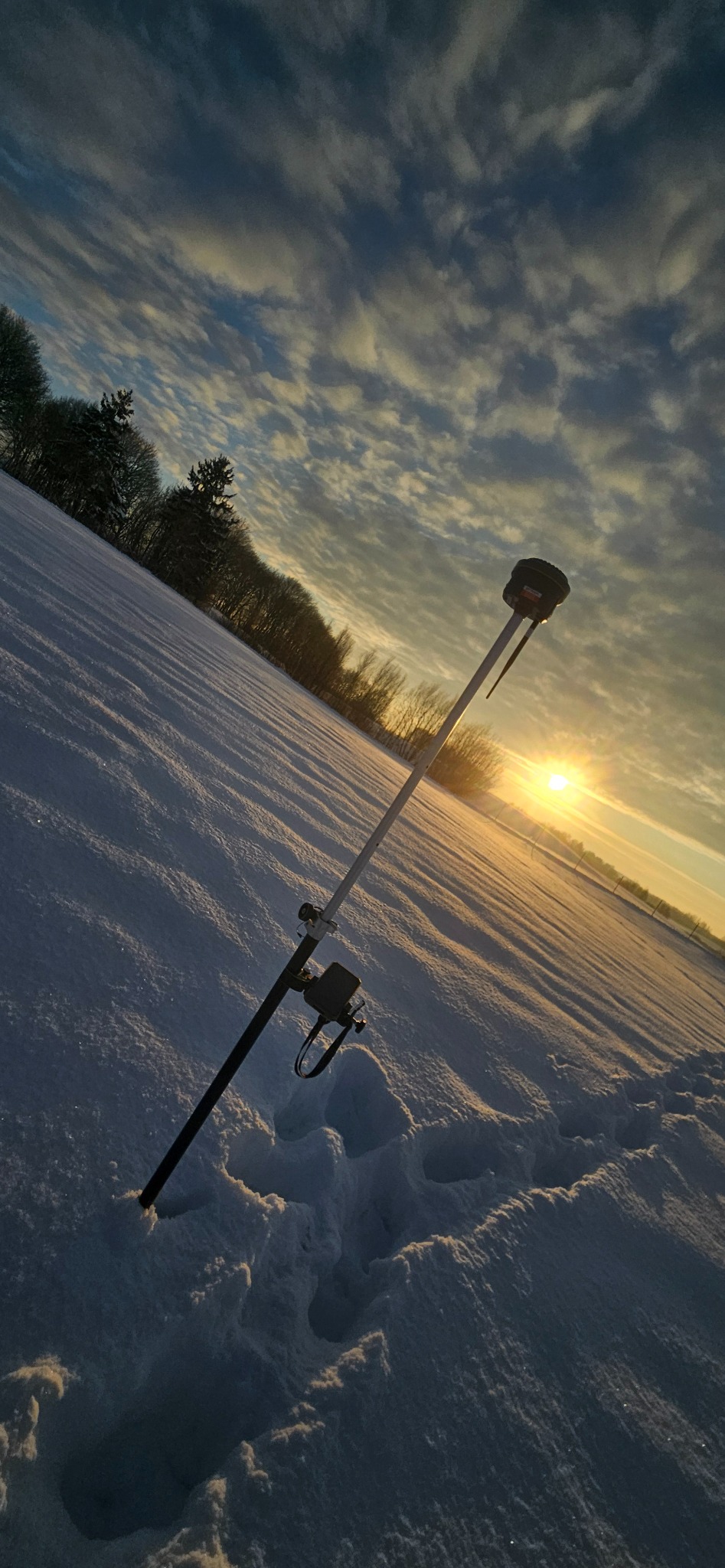 Geodezyjny odbiornik GNSS na tyczce w śnieżnym terenie oświetlonym zachodzącym słońcem. Widoczne ślady stóp w śniegu i malownicze chmury na niebie.