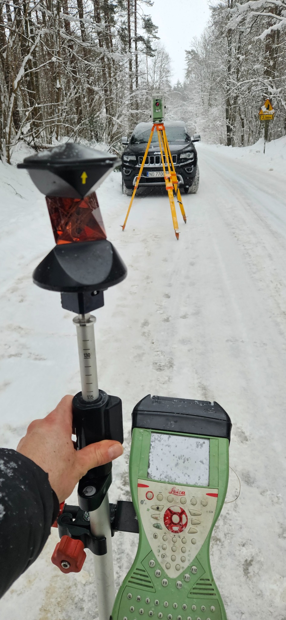 Praca w terenie zimą: tyczka geodezyjna z pryzmatem i rejestratorem Leica na tle zasypanej śniegiem drogi i samochodu Jeep z żółtymi znakami ostrzegawczymi.