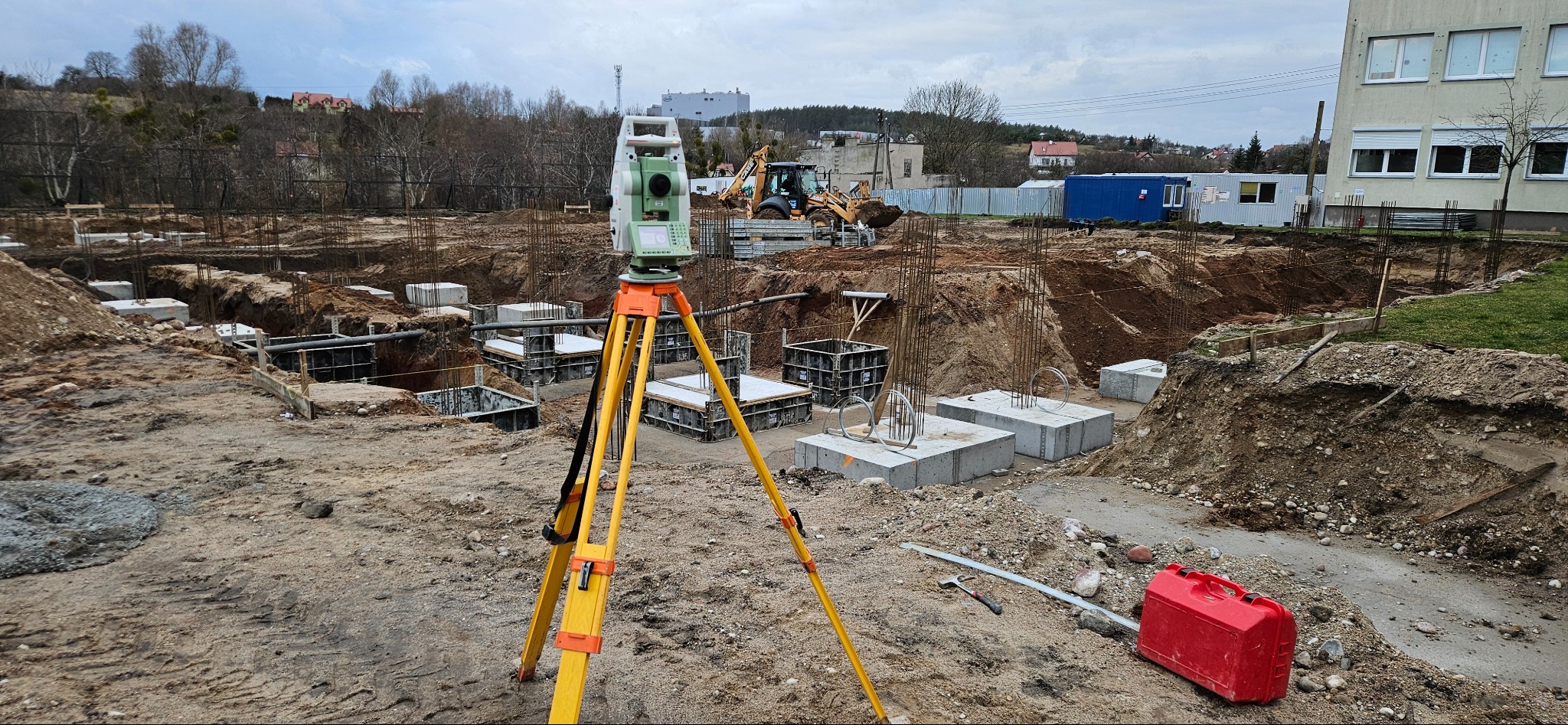 Prace ziemne pod budowę: fundamenty, zbrojenia i niwelator na żółtym statywie. W tle koparka i budynek. Widok na plac budowy w trakcie realizacji.