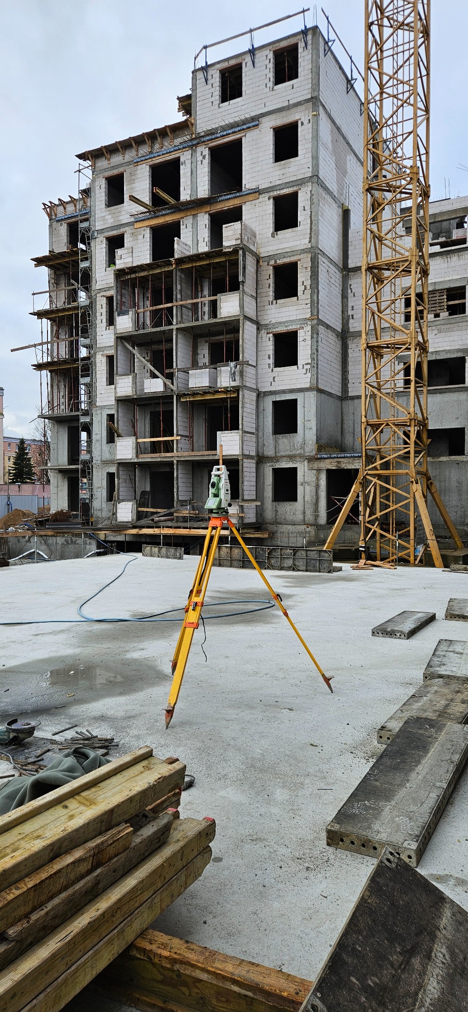 Żółty niwelator na trójnogu na tle surowego budynku w trakcie budowy i żurawia. Widoczne drewniane elementy szalunkowe i betonowe płyty.