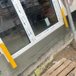 Nowe okno PCV z białymi ramami, zamontowane w ścianie z szarym tynkiem, zabezpieczone żółtymi klinami, widoczne rusztowanie i paleta w tle.