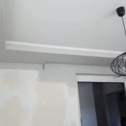 Ściana w trakcie przygotowania do malowania, widoczne ślady po gładzi, sufit z ozdobnym podwieszeniem, nowoczesna lampa wisząca z żarówką Edisona.