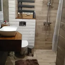 Wykończona łazienka z drewnopodobnymi płytkami, grzejnikiem drabinkowym w kolorze grafitowym, umywalką nablatową na drewnianym blacie i puszystym brązowym dywanikiem.