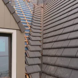 Montaż nowych, grafitowych dachówek na dachu domu jednorodzinnego, widoczne narzędzia i fragment okna.