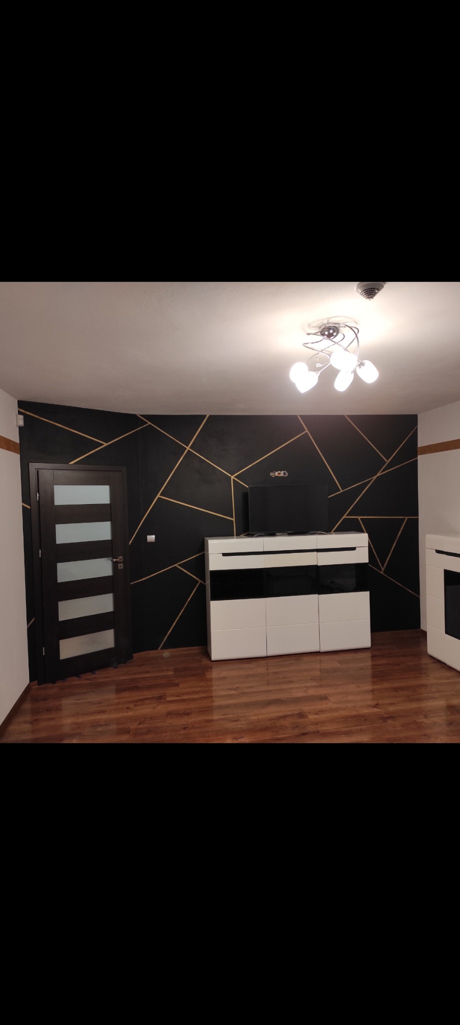 Pokój po remoncie z czarną ścianą dekorowaną złotymi liniami, brązową podłogą, białymi meblami i ciemnymi drzwiami z szybami.