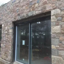 Ściana zewnętrzna budynku wykończona nieregularnym kamieniem elewacyjnym w odcieniach brązu i szarości, z widocznymi czarnymi ramami okiennymi i przewodami elektrycznymi zwisającymi wzdłuż ściany.