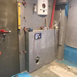 Szare płytki ceramiczne układane na ścianie w łazience, widoczne narzędzia glazurnicze, poziomica, system poziomowania płytek, instalacja podtynkowa WC w trakcie montażu, zabezpieczenie podłogi...
