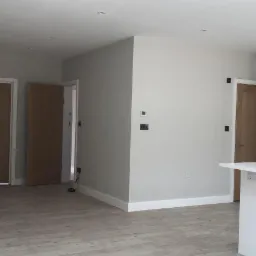 Jasne wnętrze po remoncie: szare ściany, drewniane drzwi, podłoga z paneli imitujących drewno, biała zabudowa kuchenna z wyspą.