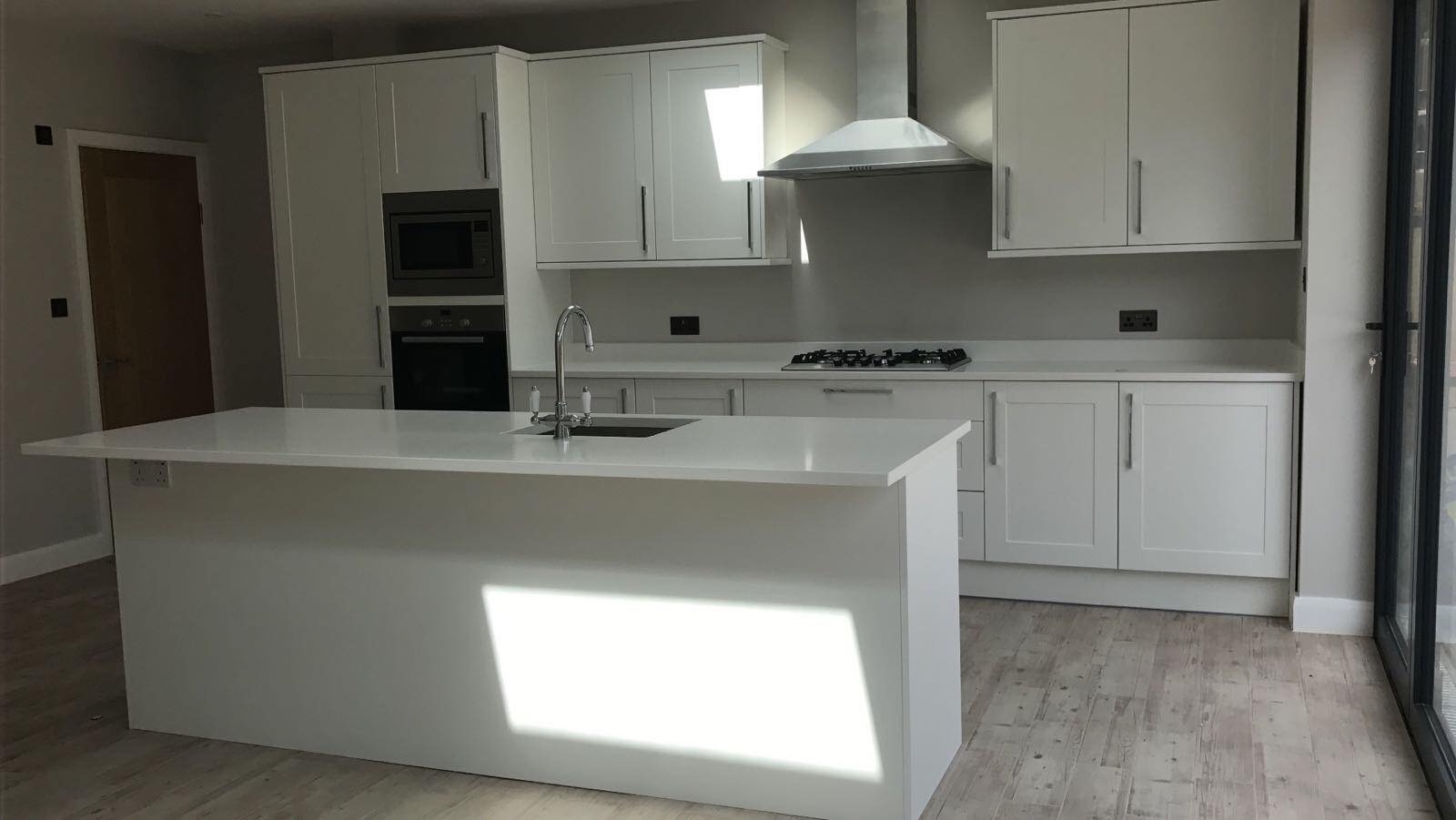 Jasna, nowoczesna kuchnia z wyspą, białe szafki z chromowanymi uchwytami, stalowy okap, drewnopodobne panele podłogowe i duże okno balkonowe w tle.