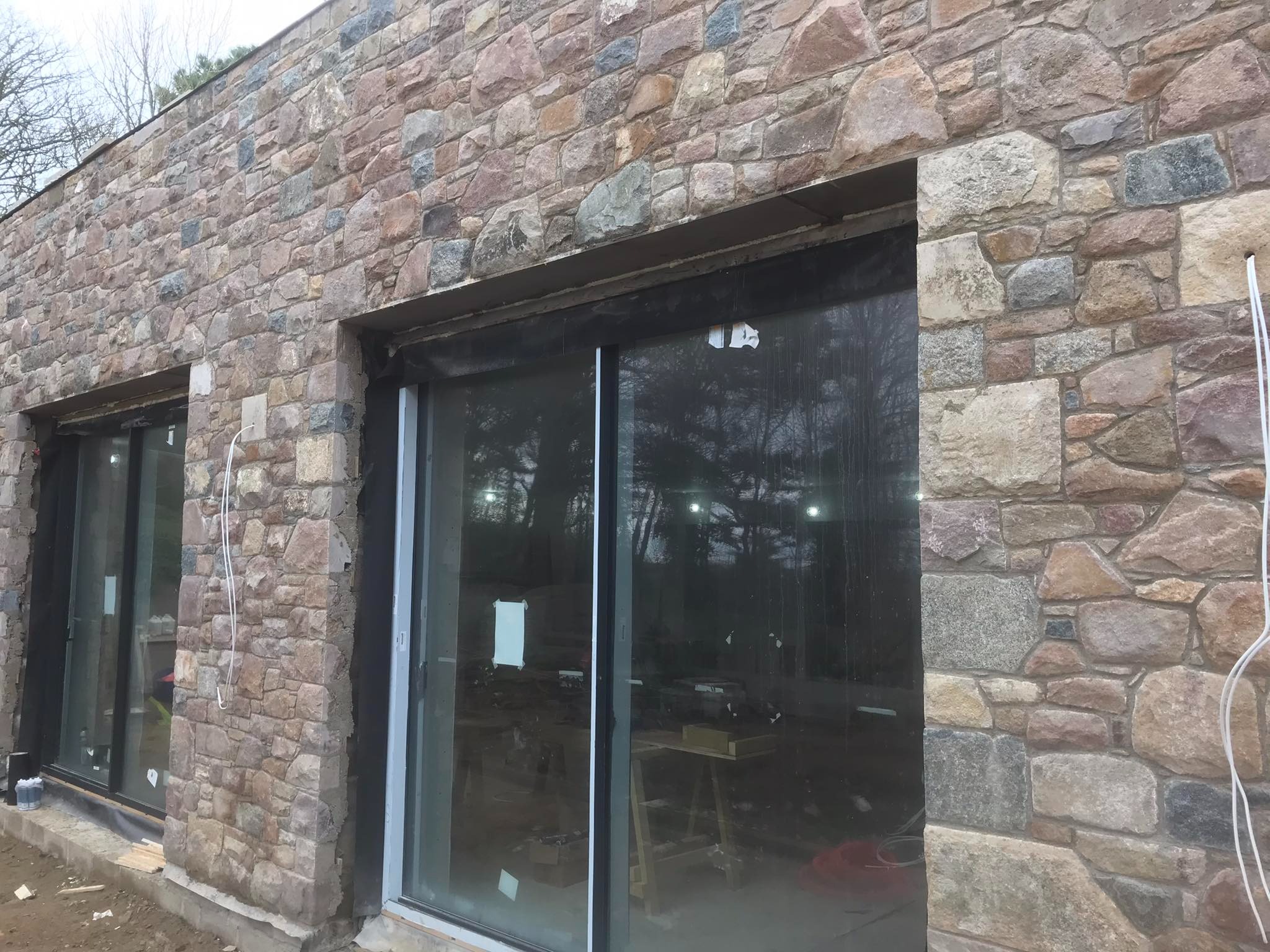 Ściana zewnętrzna budynku wykończona nieregularnym kamieniem elewacyjnym w odcieniach brązu i szarości, z widocznymi czarnymi ramami okiennymi i przewodami elektrycznymi zwisającymi wzdłuż ściany.