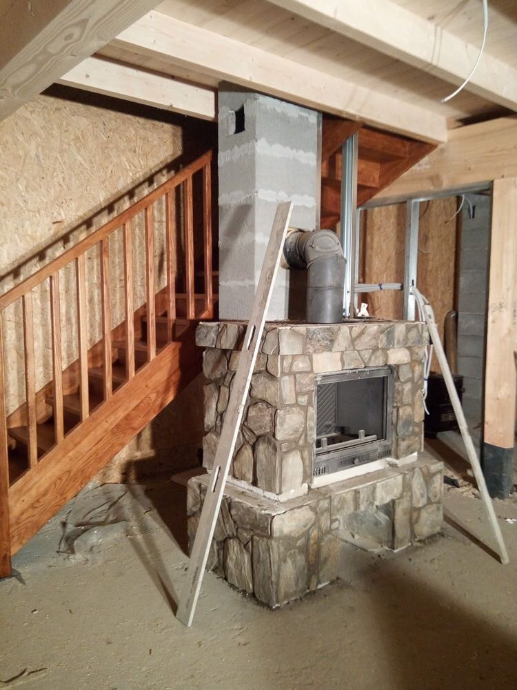 Wnętrze w trakcie wykańczania: murowany kominek obłożony kamieniem, z podłączonym metalowym wkładem kominowym, surowe drewniane schody i poddasze z widoczną konstrukcją stropu, poziomica oparta...