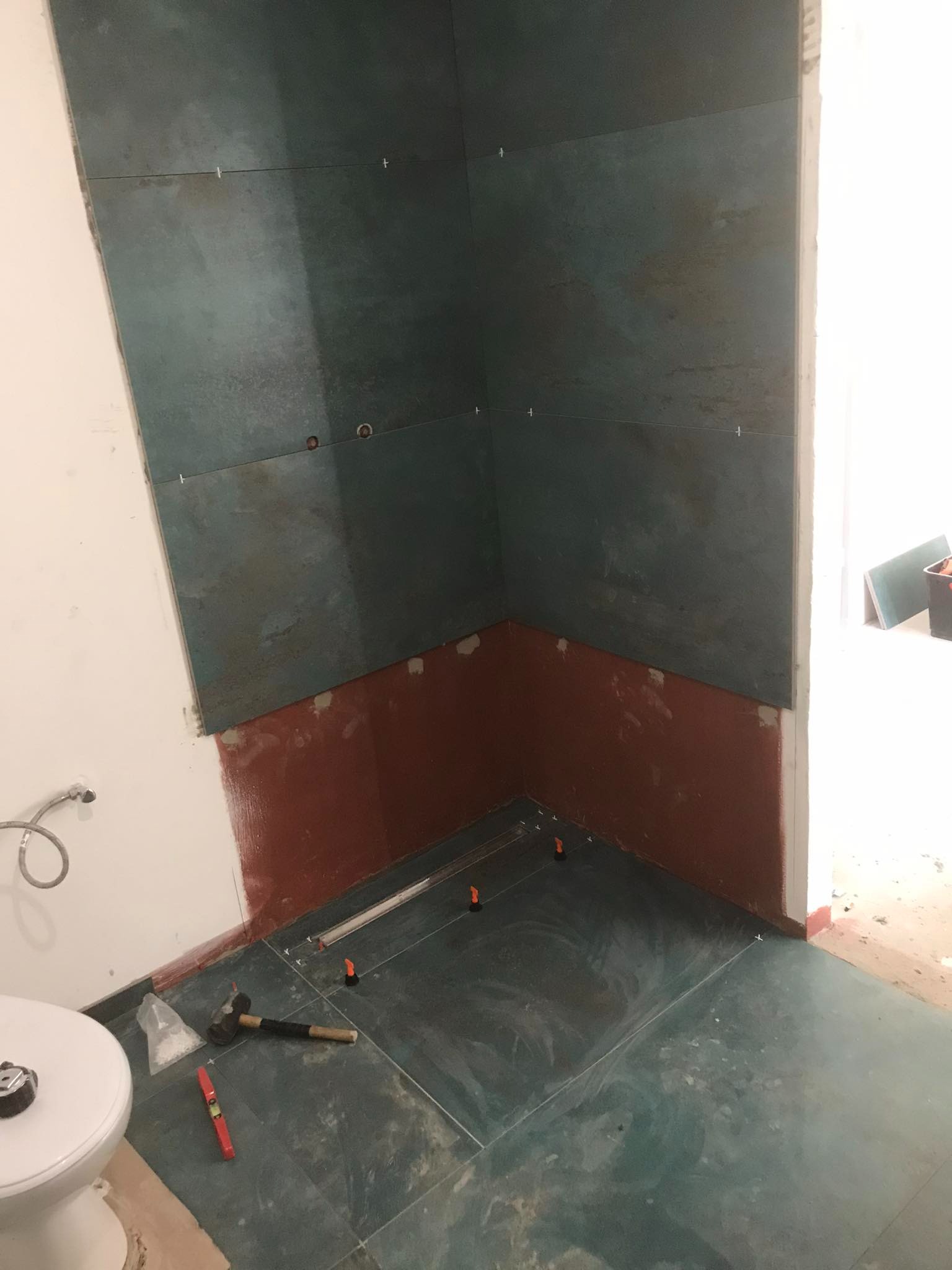 W trakcie prac: ciemnozielone płytki imitujące kamień na ścianach i podłodze łazienki, widoczne poziomica, gumowy młotek i system poziomowania płytek.