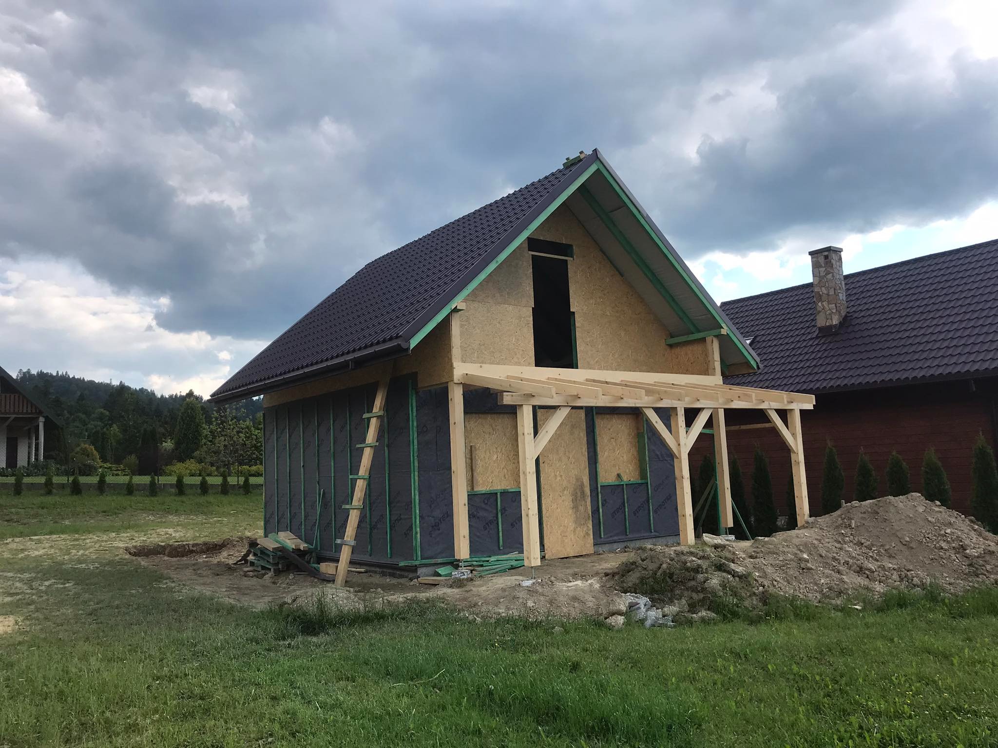 Surowy stan budowy domu z drewnianą konstrukcją szkieletową, czarną membraną izolacyjną i częściowo ukończonym dachem z metalowej blachy, widoczna drabina oparta o ścianę i drewniany szkielet...