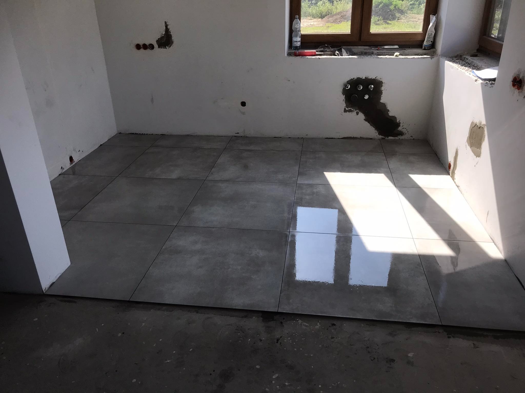 Świeżo ułożone, duże, szare płytki podłogowe w trakcie prac wykończeniowych w pomieszczeniu z oknem, widoczne gniazdka elektryczne i niedokończone ściany.