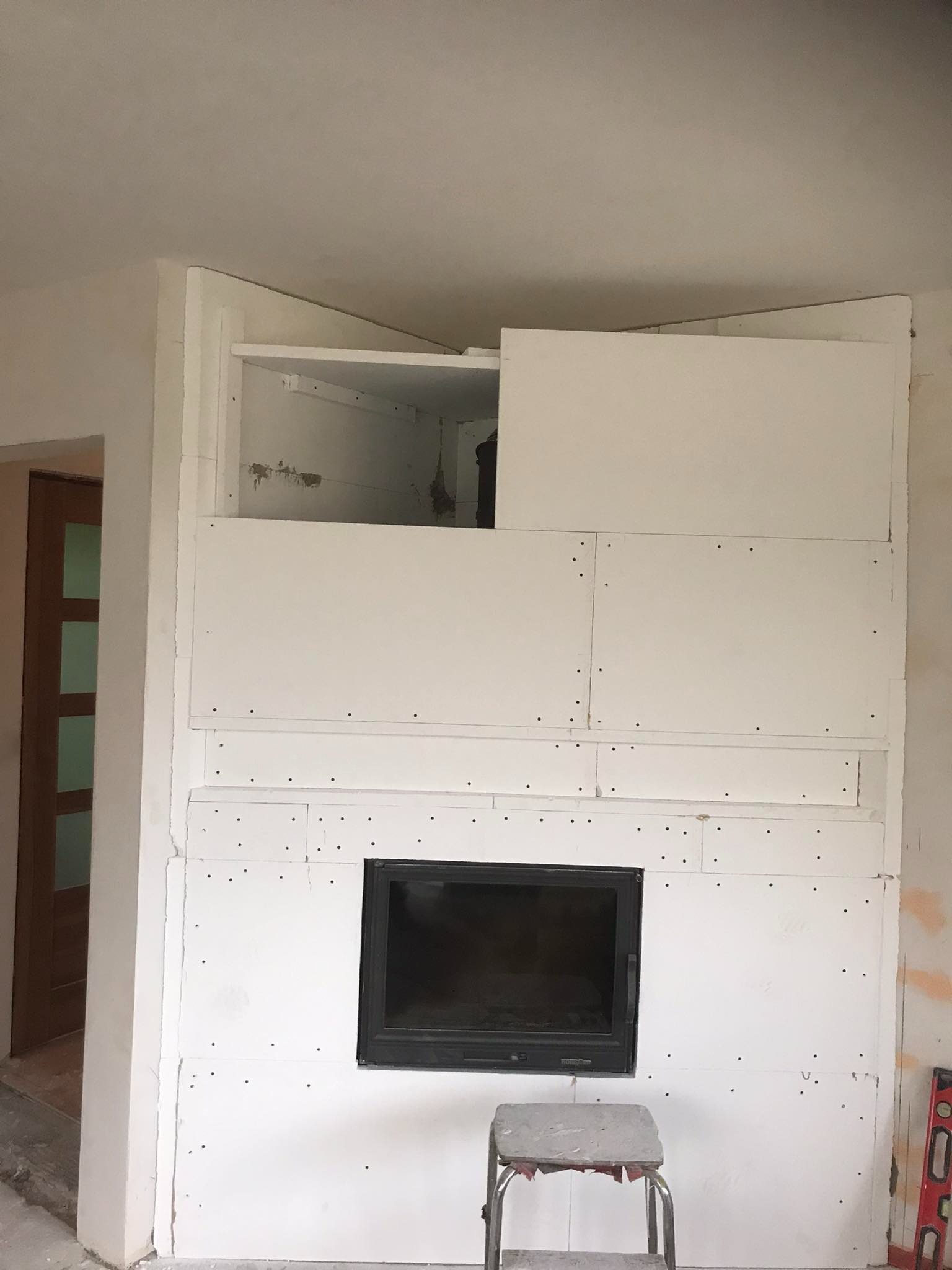 Konstrukcja obudowy kominka z płyt kartonowo-gipsowych w trakcie montażu, widoczne łączenia płyt i otwór na wkład kominkowy, drabinka i poziomica w kadrze.