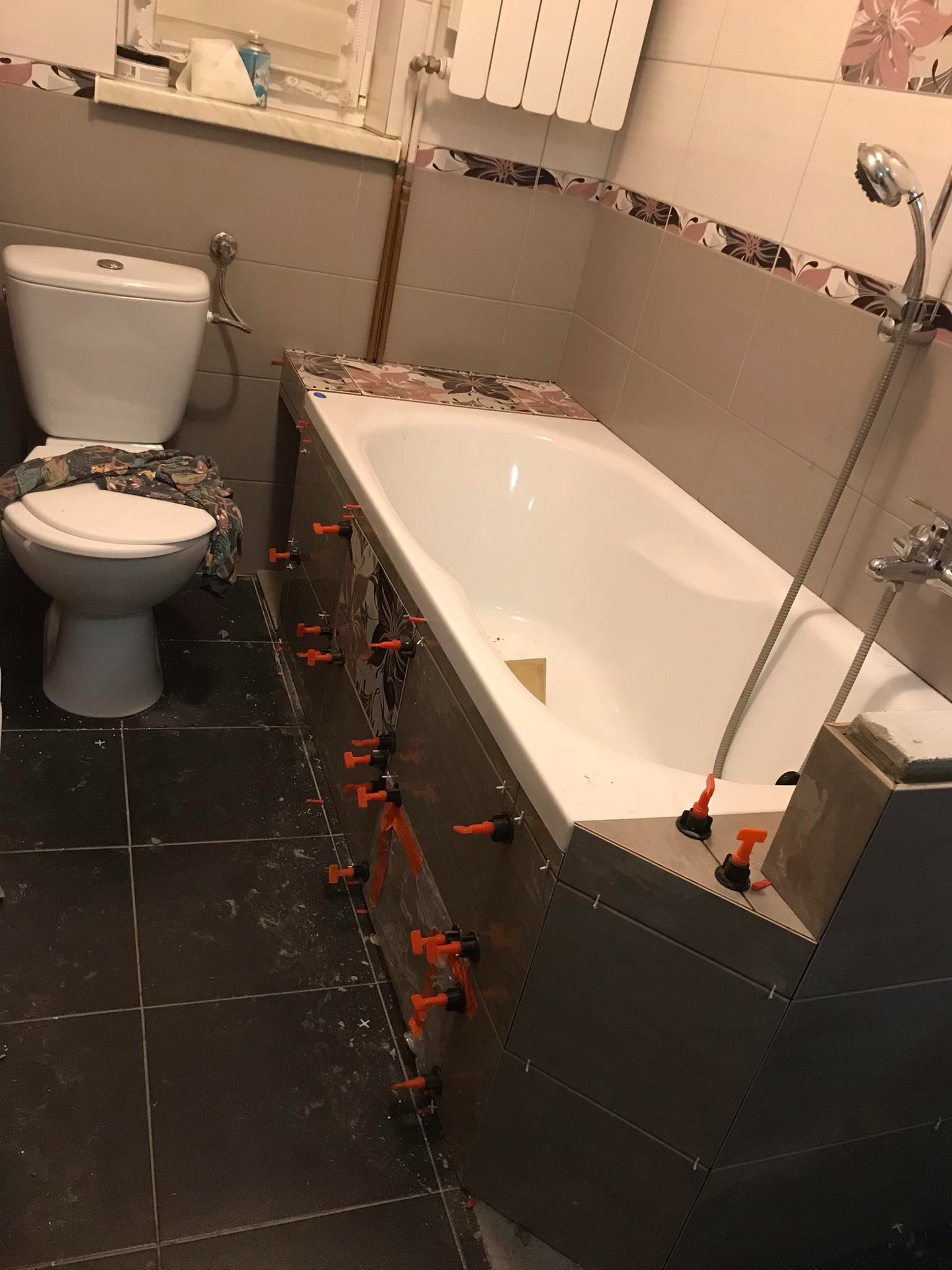 Montaż płytek na obudowie wanny w łazience w Polańczyku, widoczne pomarańczowe elementy poziomujące, czarne płytki podłogowe i toaleta.