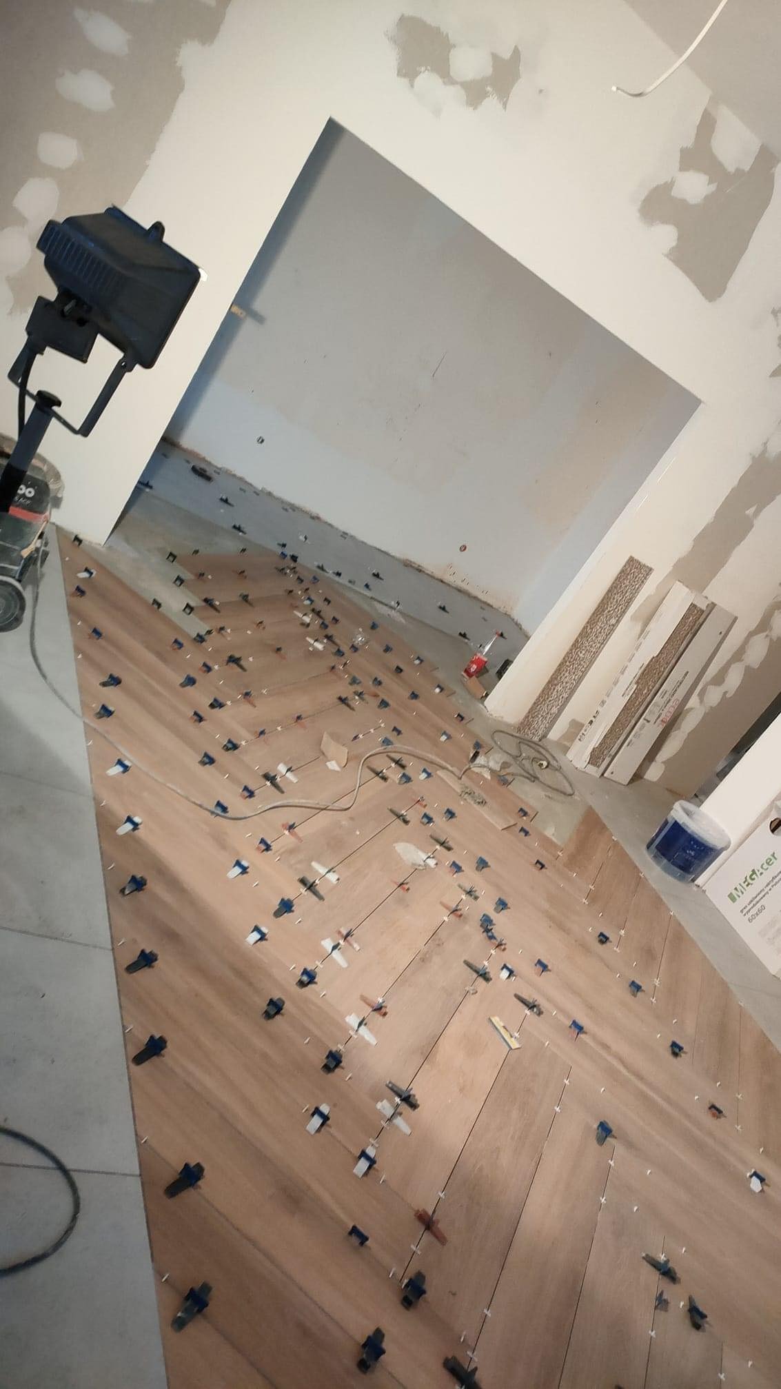 Ukośne ułożenie płytek podłogowych w trakcie prac, z użyciem systemu poziomowania, widoczne płytki pod ścianą i narzędzia pracy, ściany w stanie surowym.